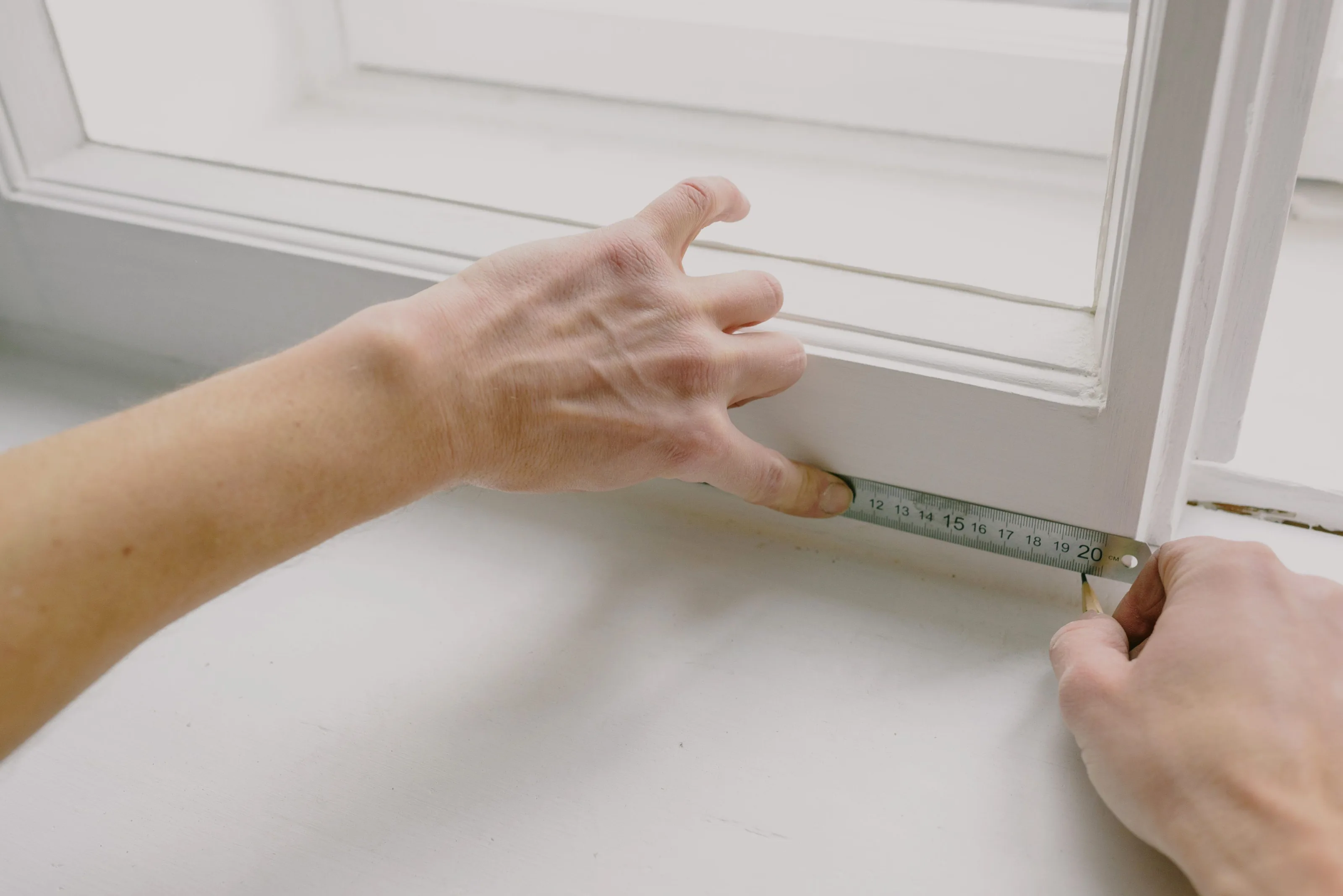 Two hands checking a white window frame with a steel ruler