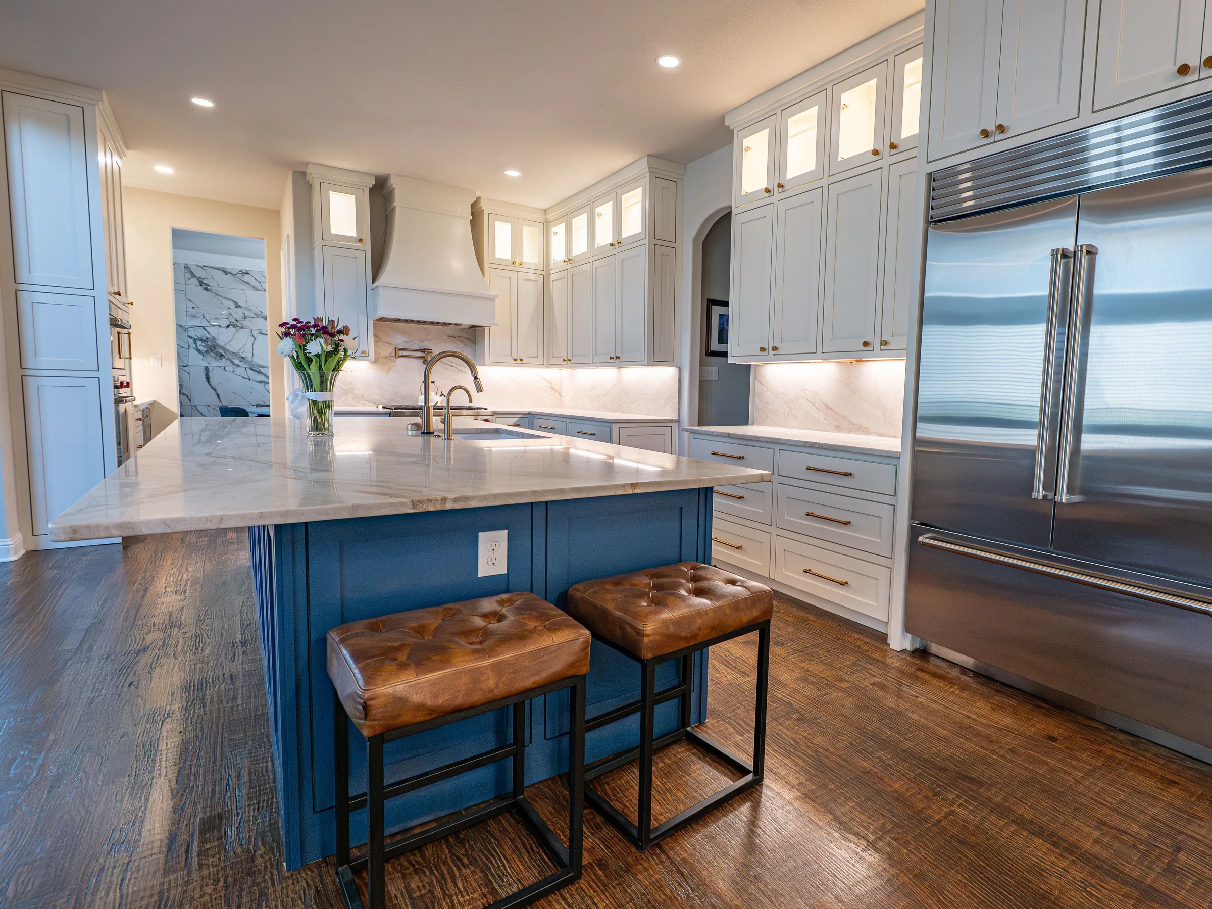 A kitchen with a navy island under a marble top, two leather stools, white cabinets and a wide steel refrigerator on wood floors