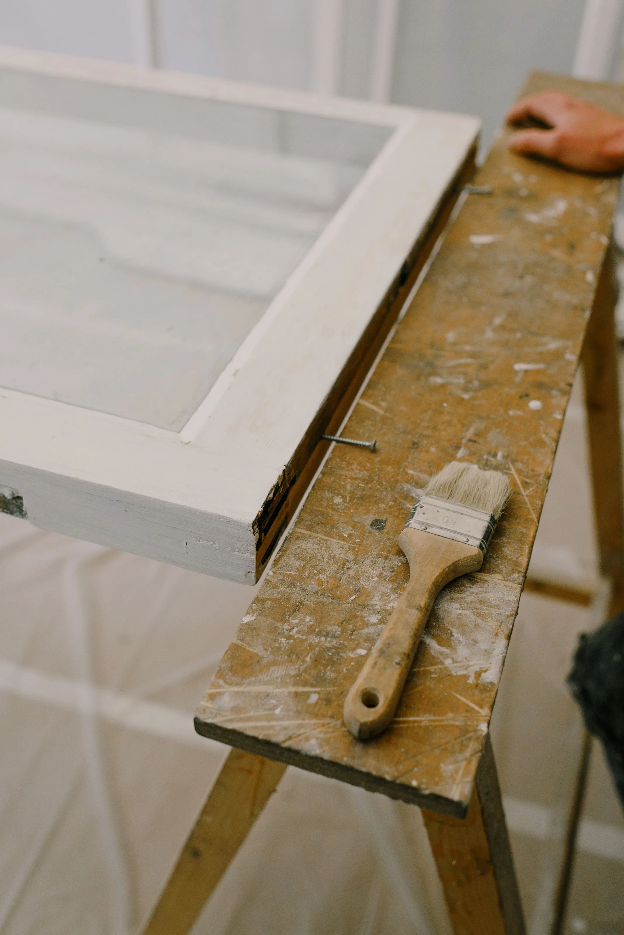 A primed window sash resting on a sawhorse beside a paintbrush, plastic sheeting on the floor below