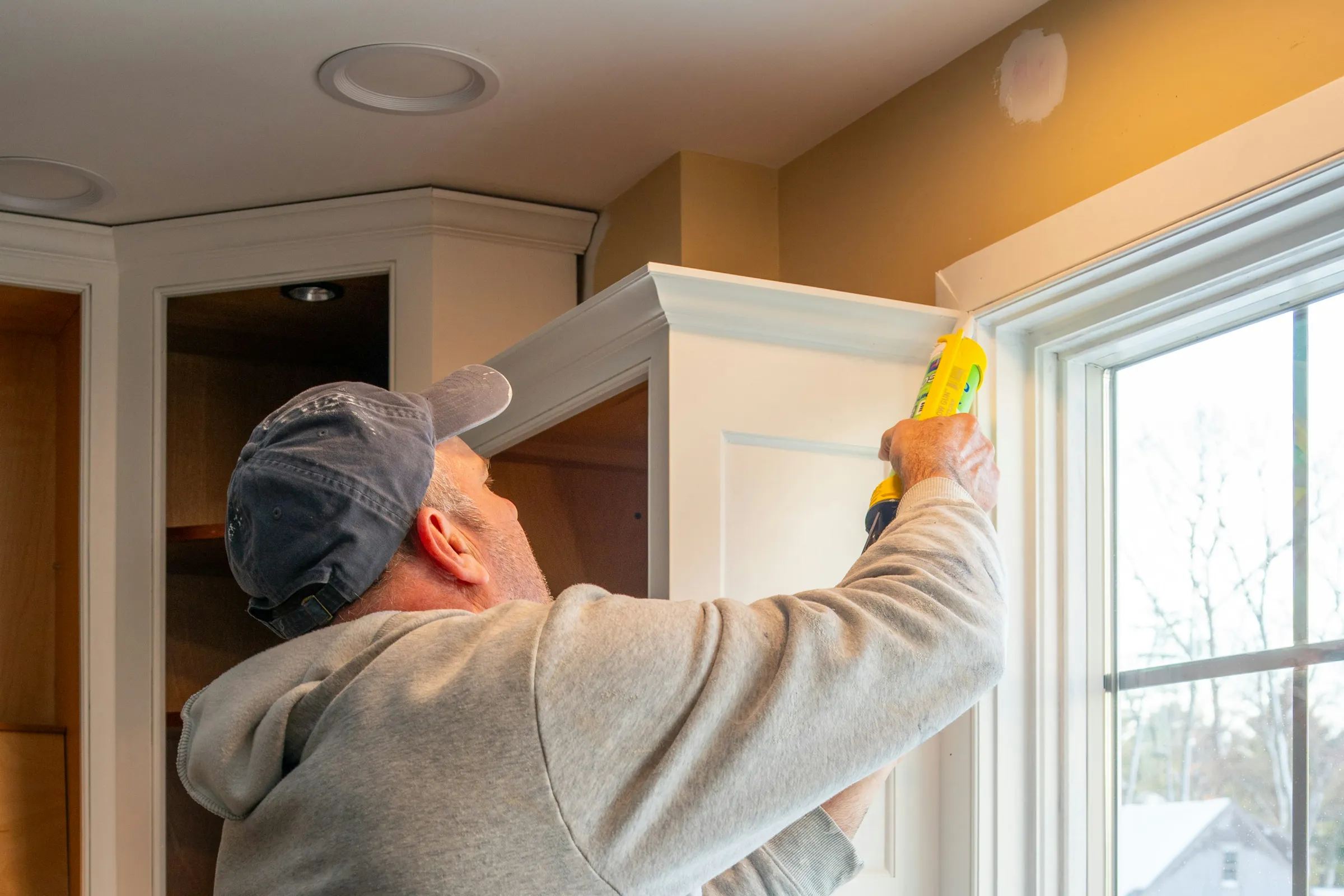 A painter seen from behind running a bead of caulk along trim beside a window, the counter under plastic