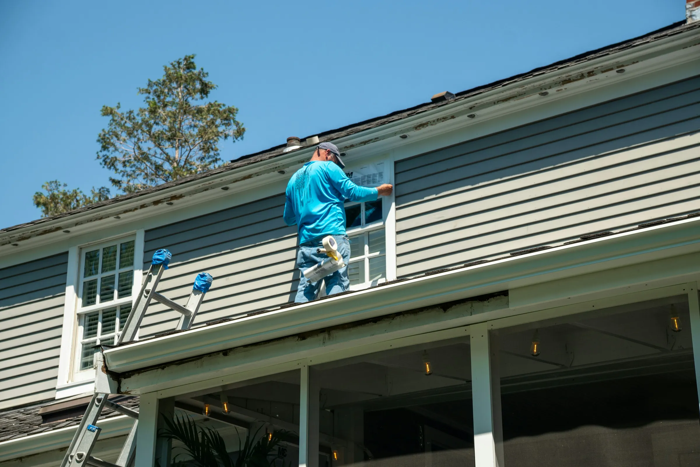 A worker on a porch roof fitting flashing around an upstairs window of a grey-sided house