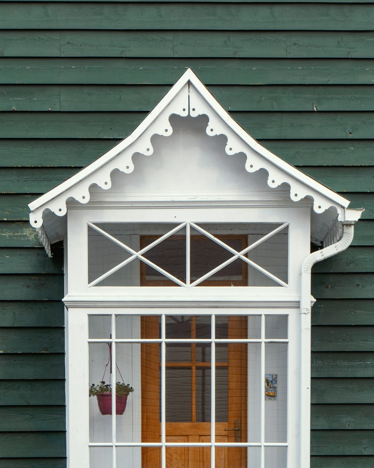 A white gable window bay with scalloped trim on dark green clapboard, a wood door behind its glass