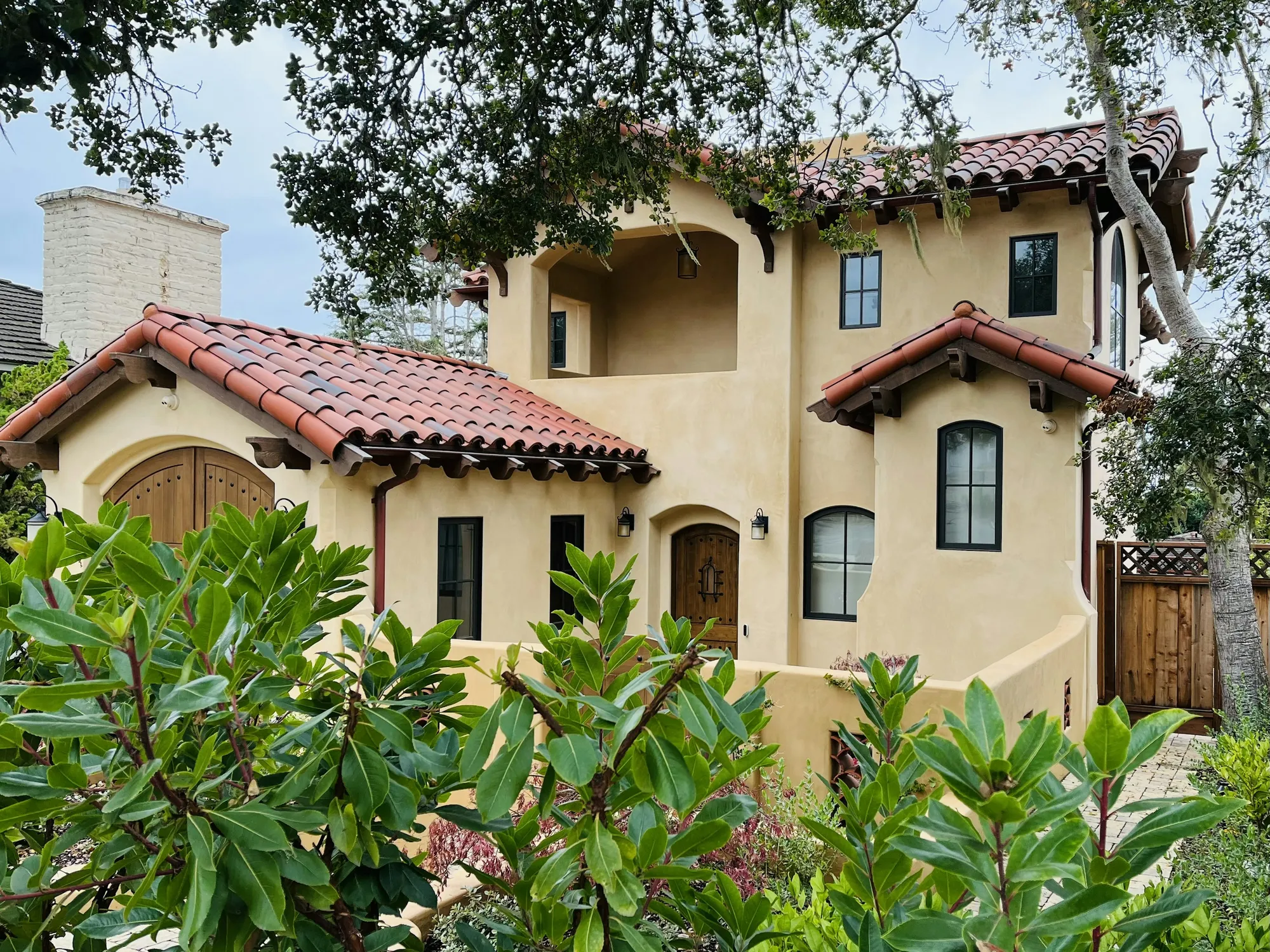 A two-storey Spanish-style stucco house with red clay tile roofs behind flowering shrubs
