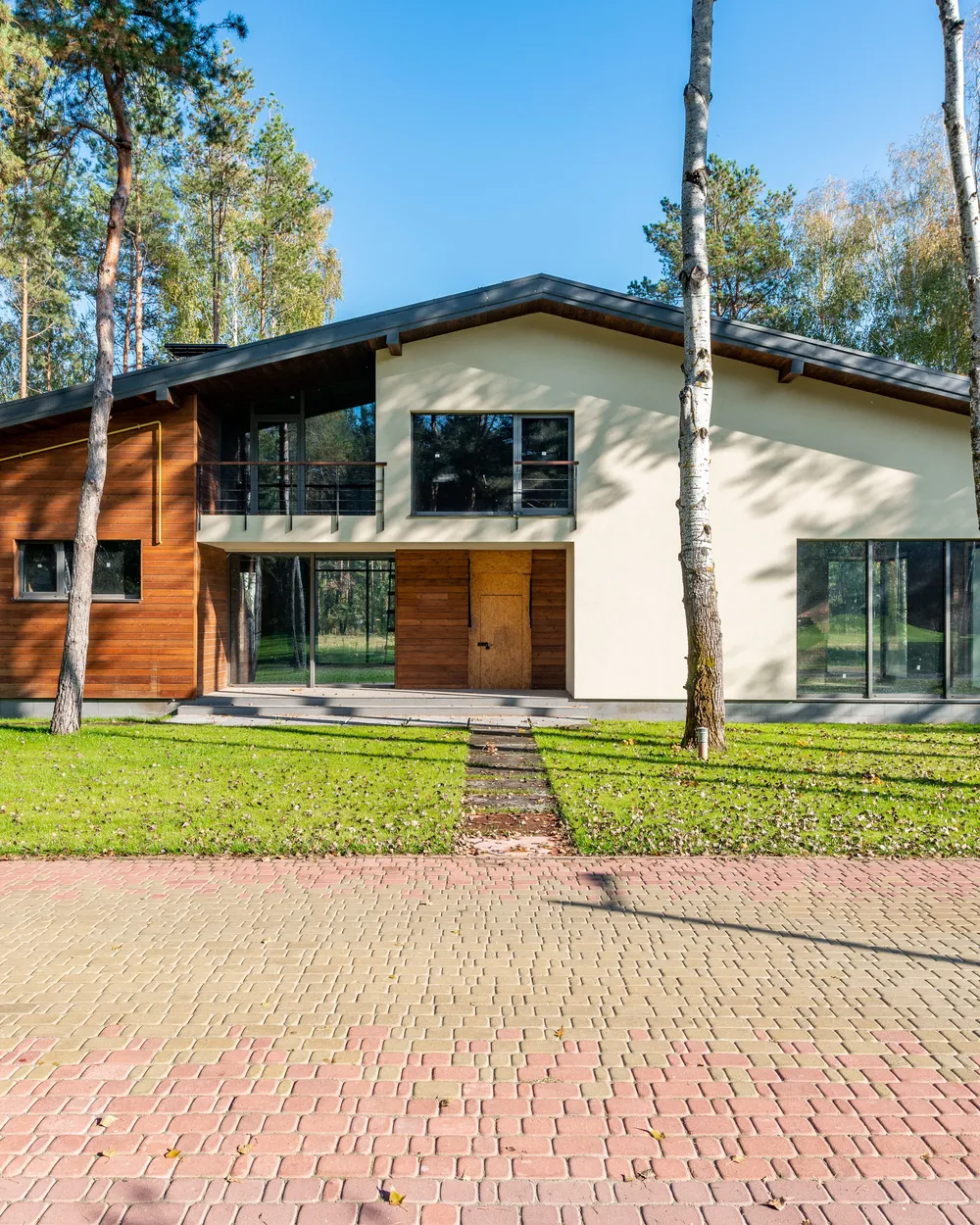 A two-storey house among tall pines, with a lawn and a wide paved forecourt in front