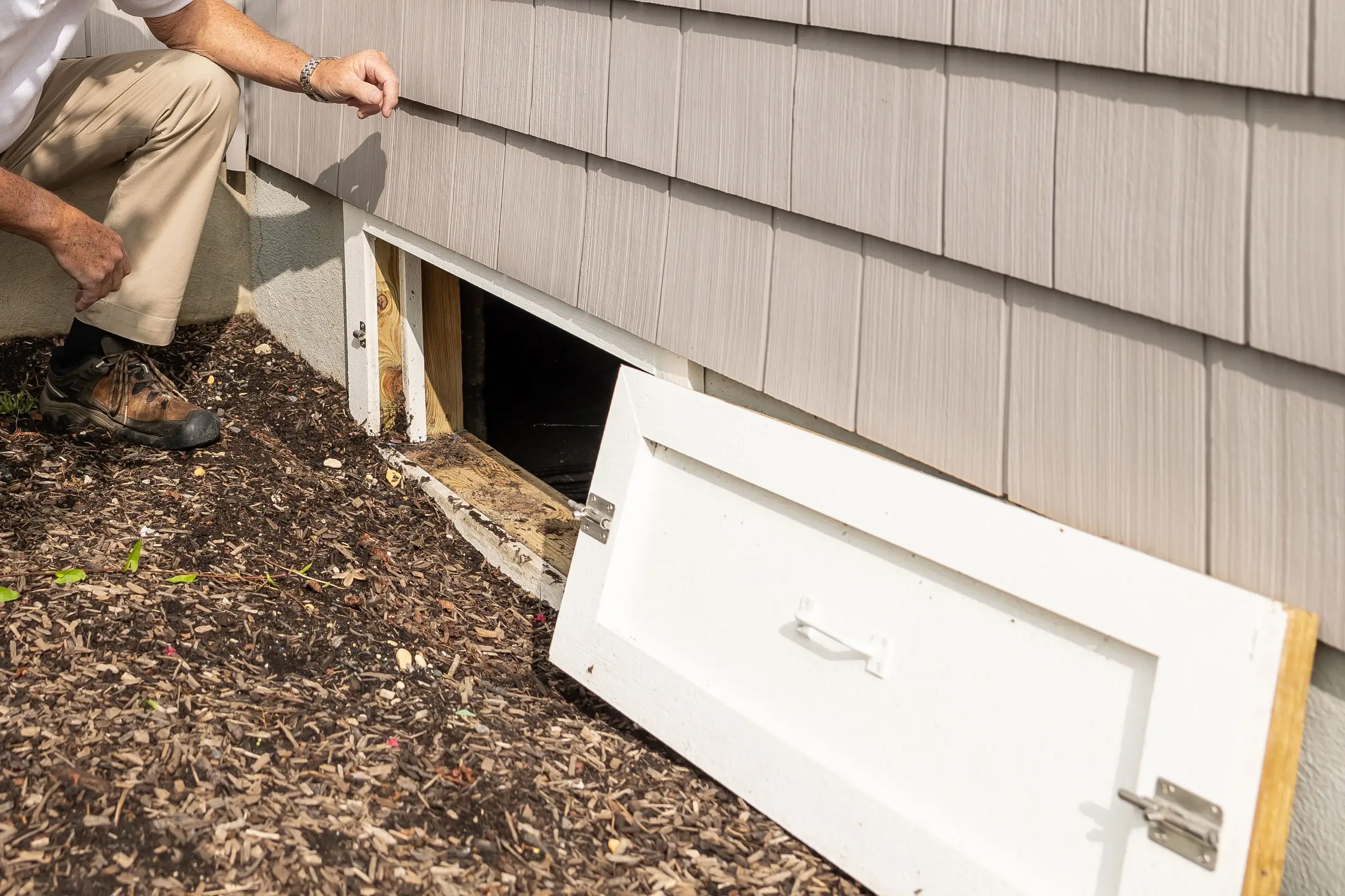 A hand rests on shingle siding beside an open crawl-space hatch at the base of a house wall