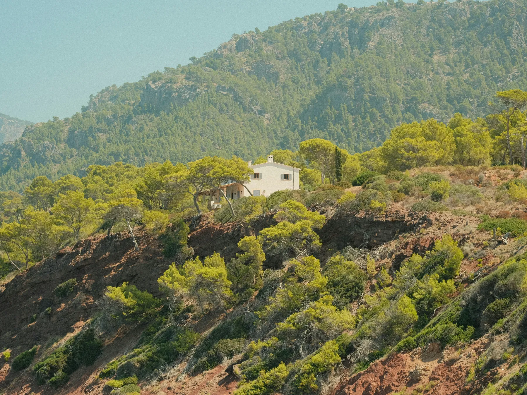 A white house on a steep hillside, surrounded by pine scrub below forested slopes